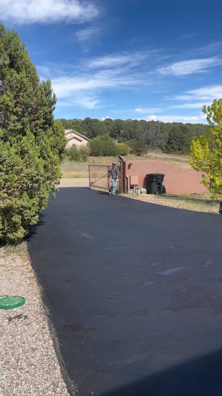 Freshly sealed black asphalt driveway leading to a gated entry in the trees
