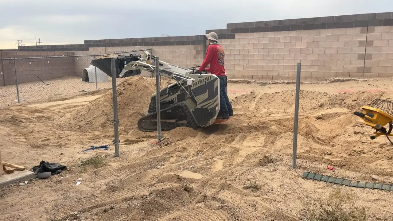 Mini track loader grading a walled backyard beside chain-link fence posts